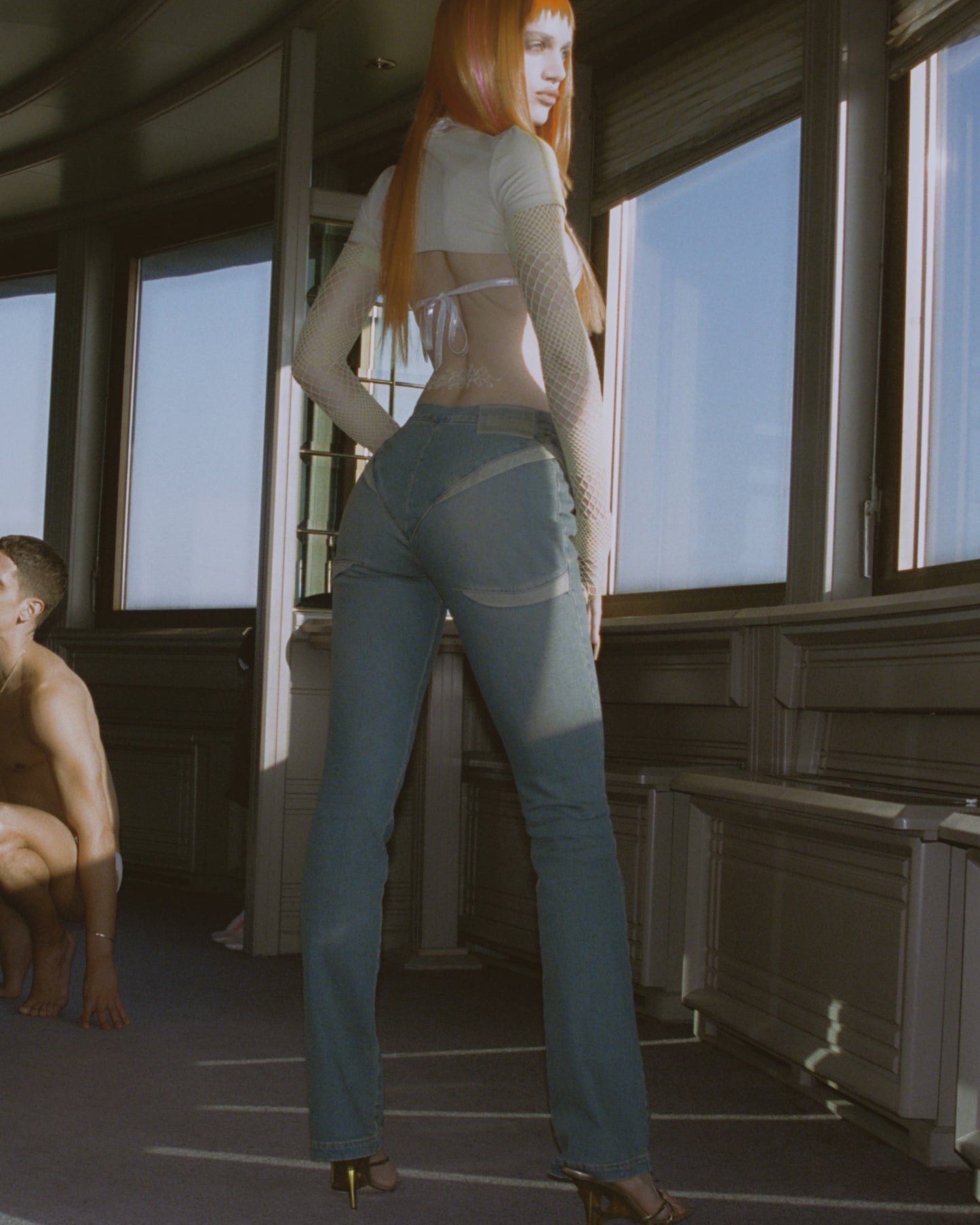 A model wearing light blue sandasimona viper denim jeans paired with the white shirt and gold shoes standing in the room with big windows and a man in the back.