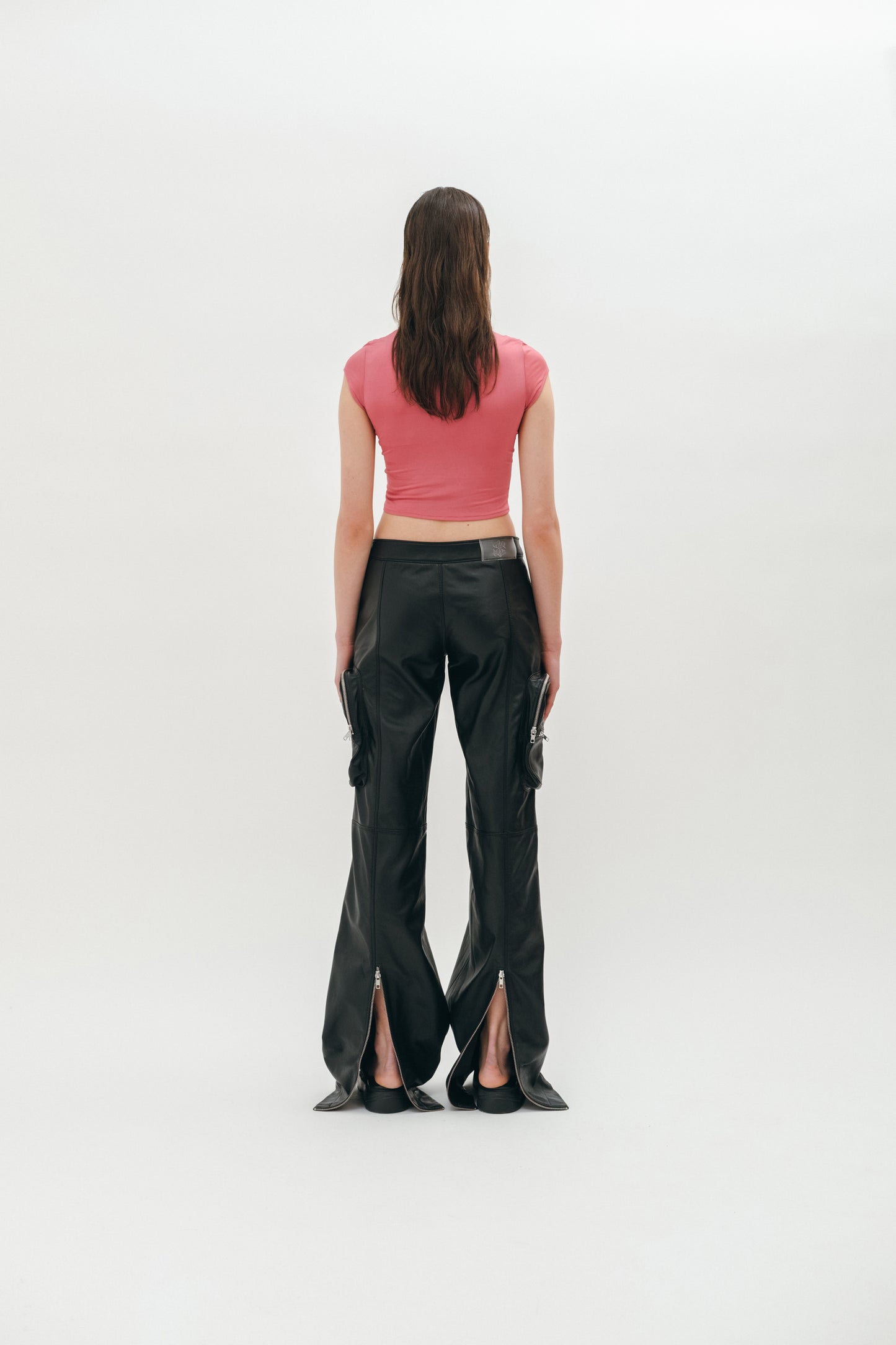 A person standing wearing black leather pants with cargo pockets, zip slits on the back, and a pink top showing the pants from the back.