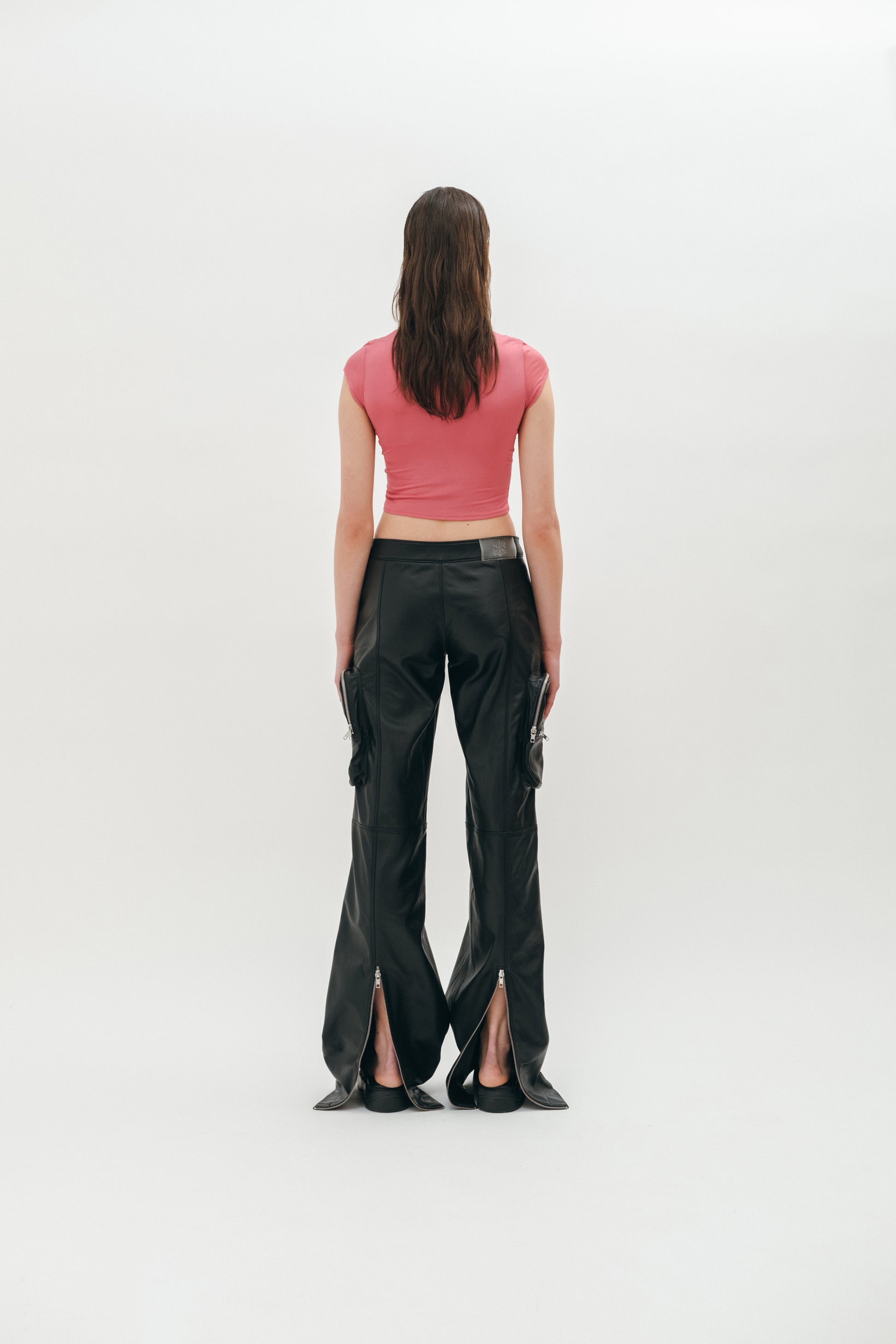 A person standing wearing black leather pants with cargo pockets, zip slits on the back, and a pink top showing the pants from the back.